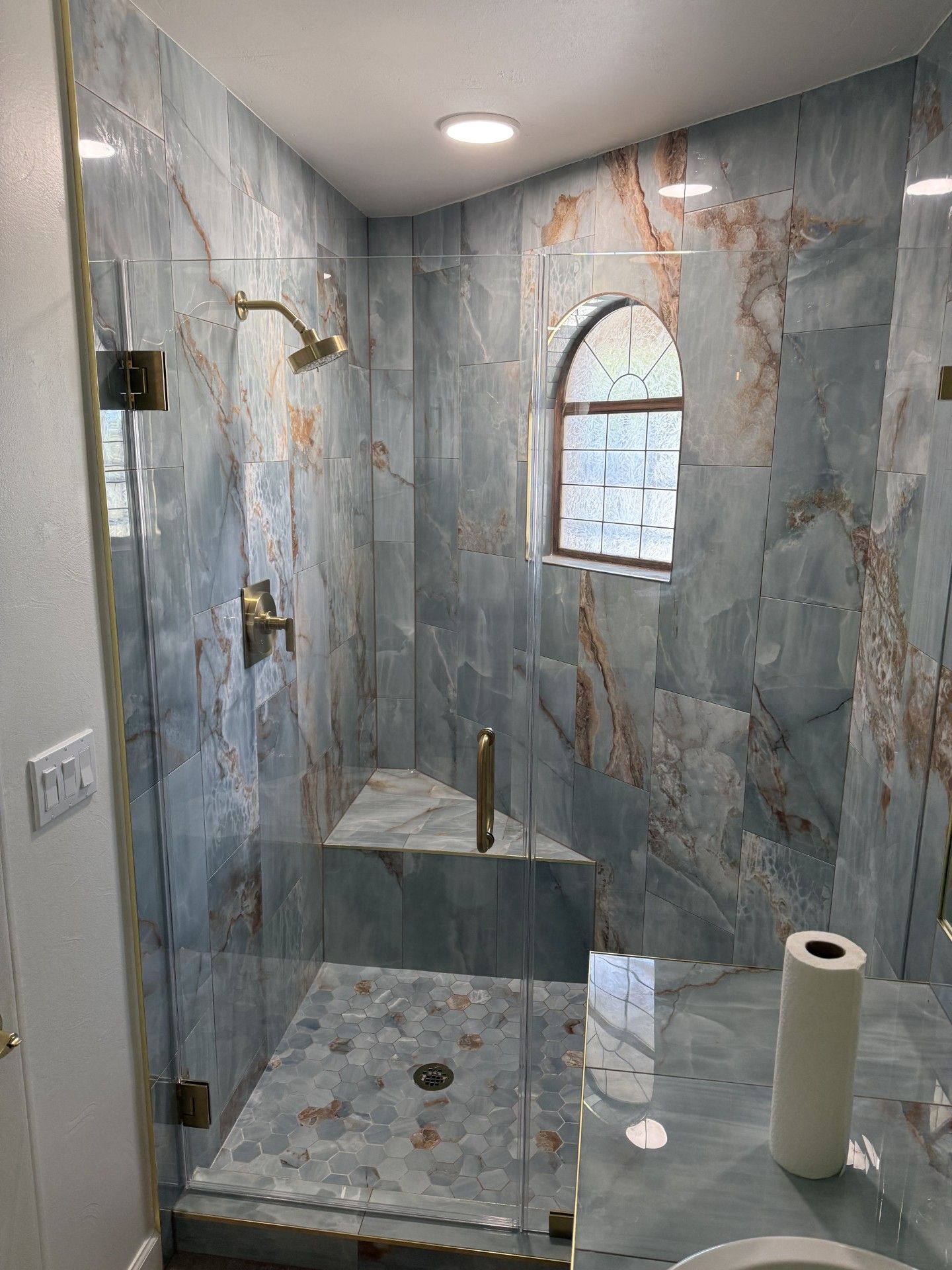 Blue marble-tiled shower with glass door, gold fixtures, and small arched window.