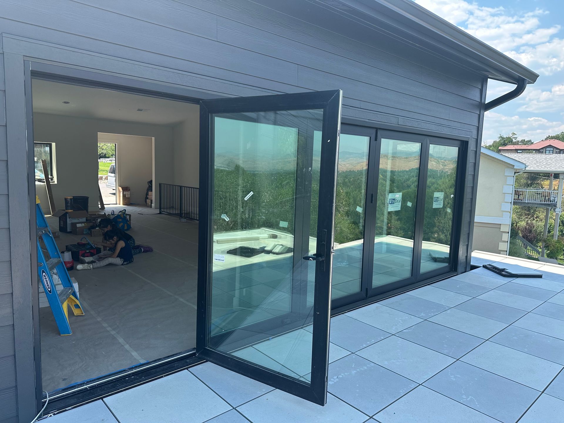 Open glass folding doors on a house, revealing interior. Gray siding, tiled patio.