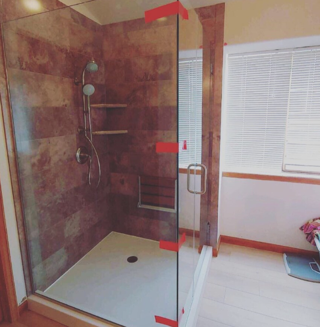 Walk-in shower with brown tile walls, glass door, and white floor. Window with blinds and a scale are nearby.