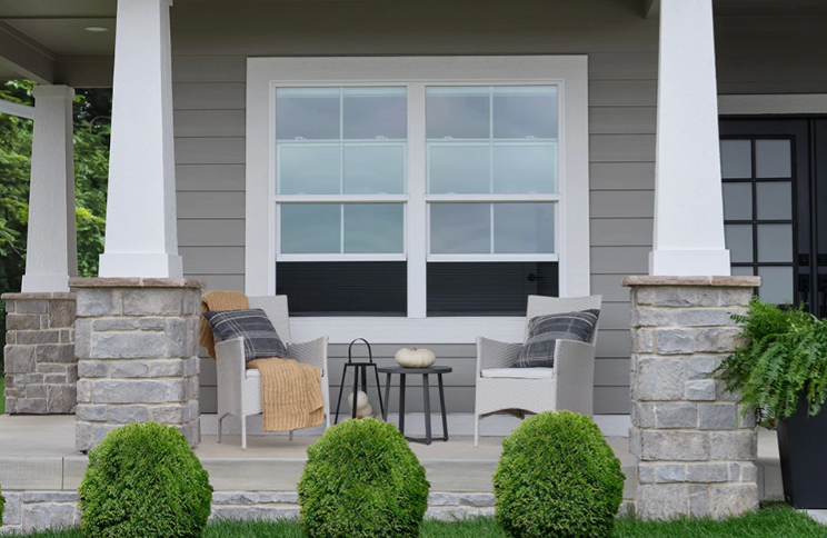 A covered front porch with white chairs, a small table, and a large window. Gray siding and stone columns.