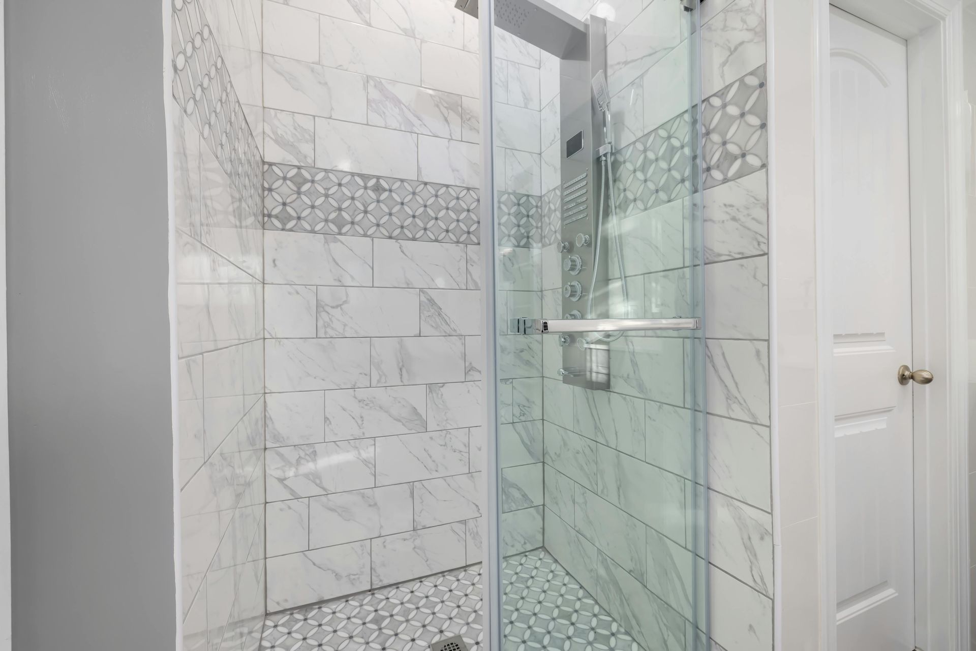 A modern, white-tiled shower with glass doors and a silver showerhead; adjacent to a white door.