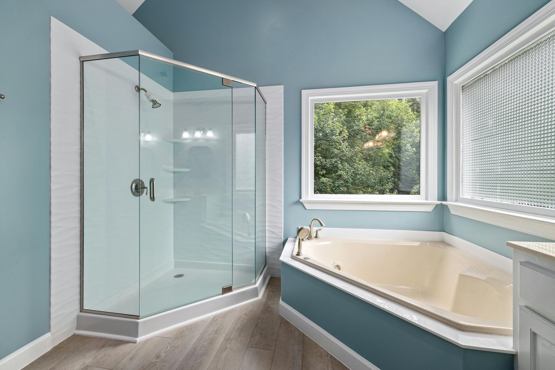 Bathroom with a corner shower, bathtub, and window; blue walls, white trim, and gray flooring.
