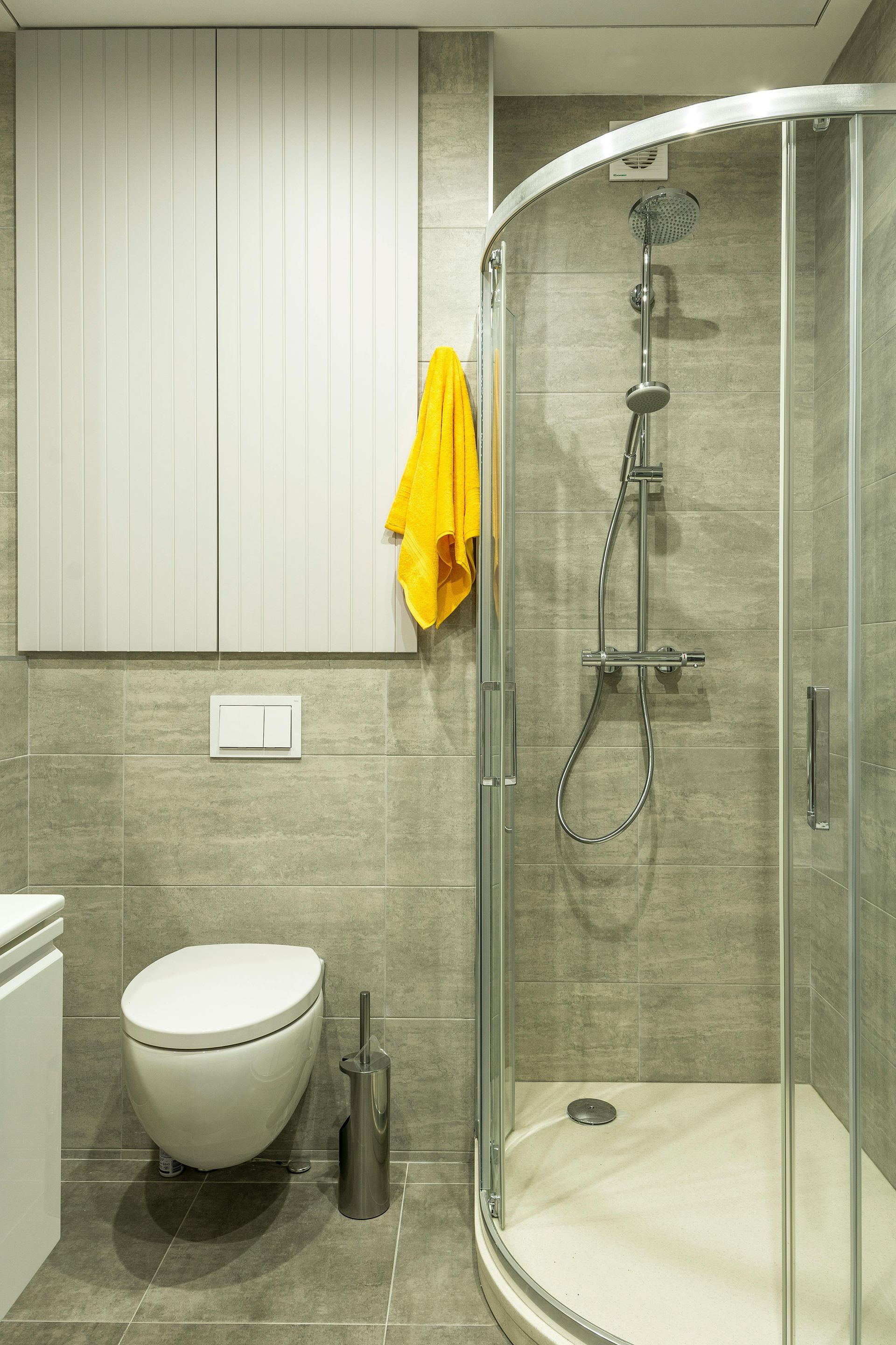 Small bathroom with shower, toilet, cabinet, and a yellow towel. Gray tile walls.