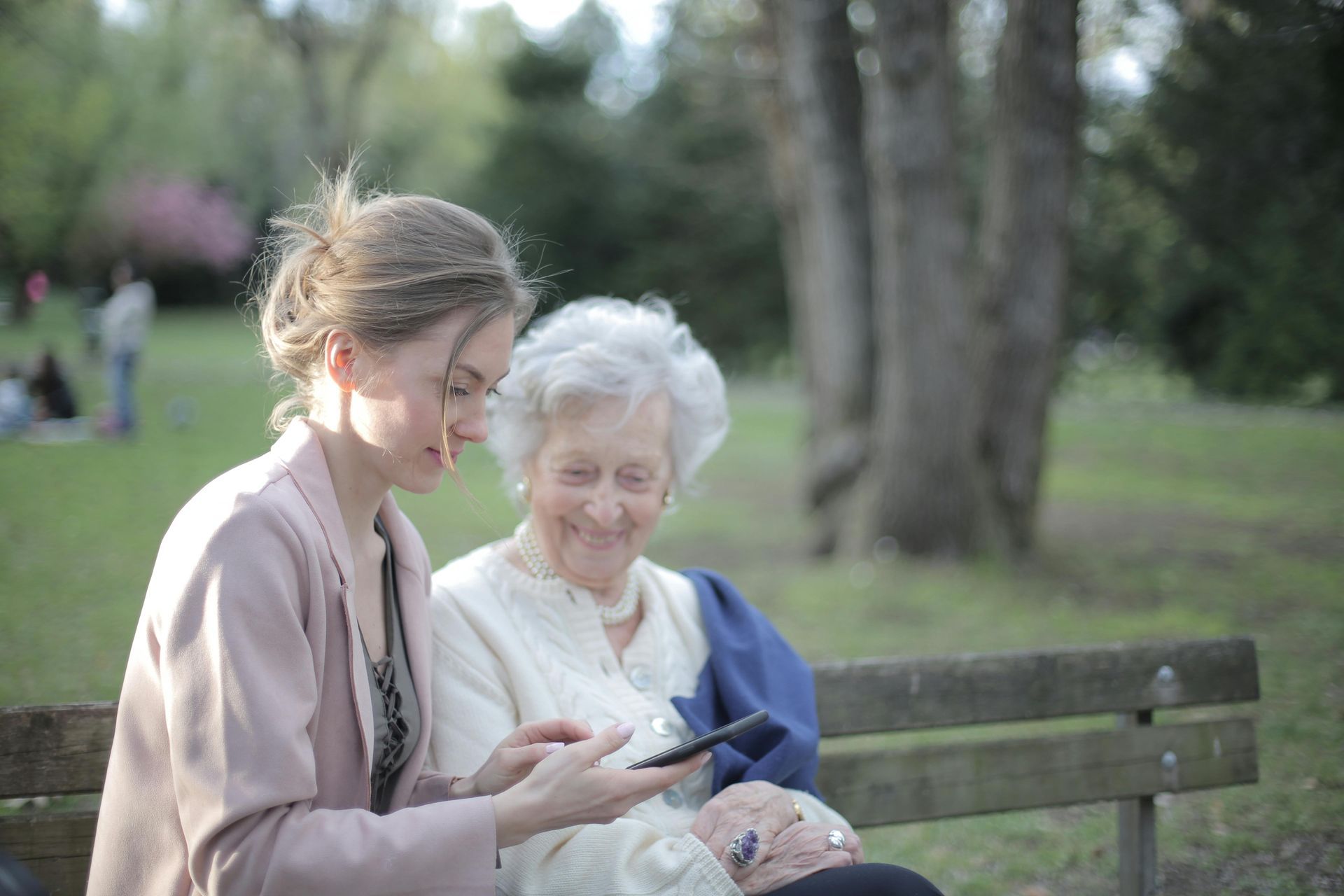 Woman showing a phone to an elderly woman on a park bench; both are smiling.