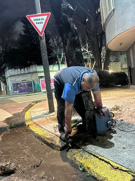 Un trabajador inclinado sobre una acera mojada, retirando escombros junto a una señal de tráfico roja y blanca por la noche.