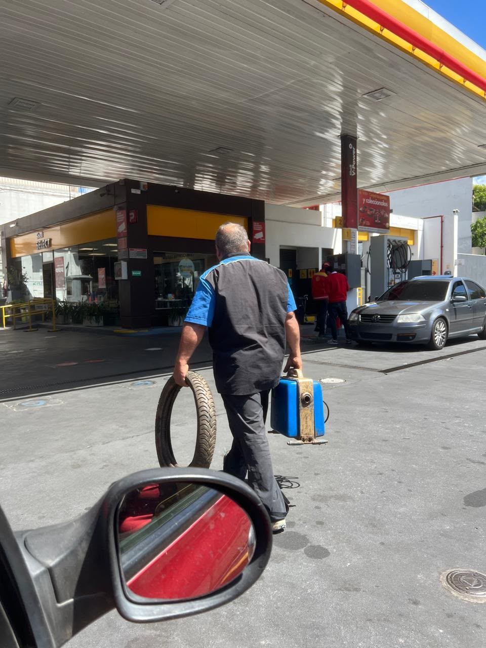 Un hombre lleva dos neumáticos por la explanada de una gasolinera, cerca de un surtidor y de un coche aparcado