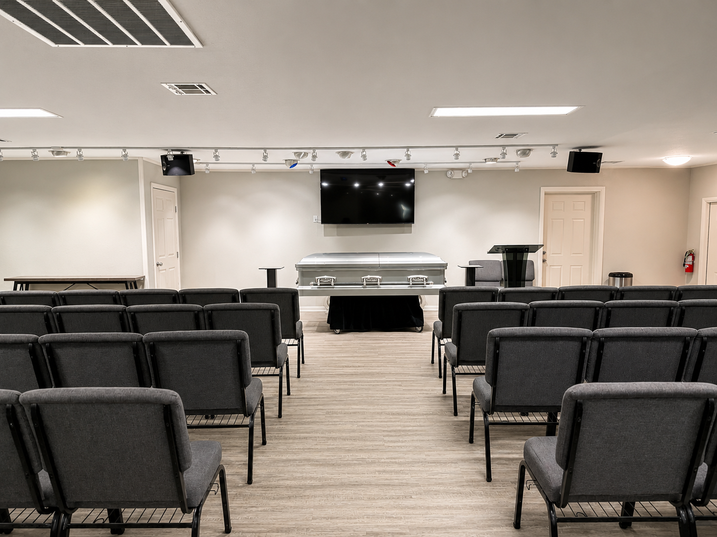 Empty room with rows of chairs facing a wall-mounted TV and podium under ceiling lights
