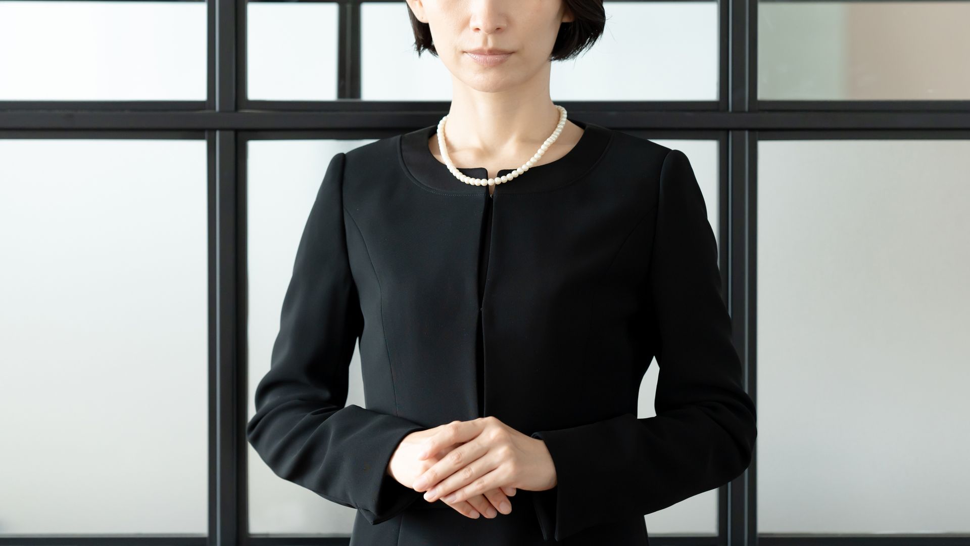 Person in a black blazer standing with hands clasped in front of a windowed wall.