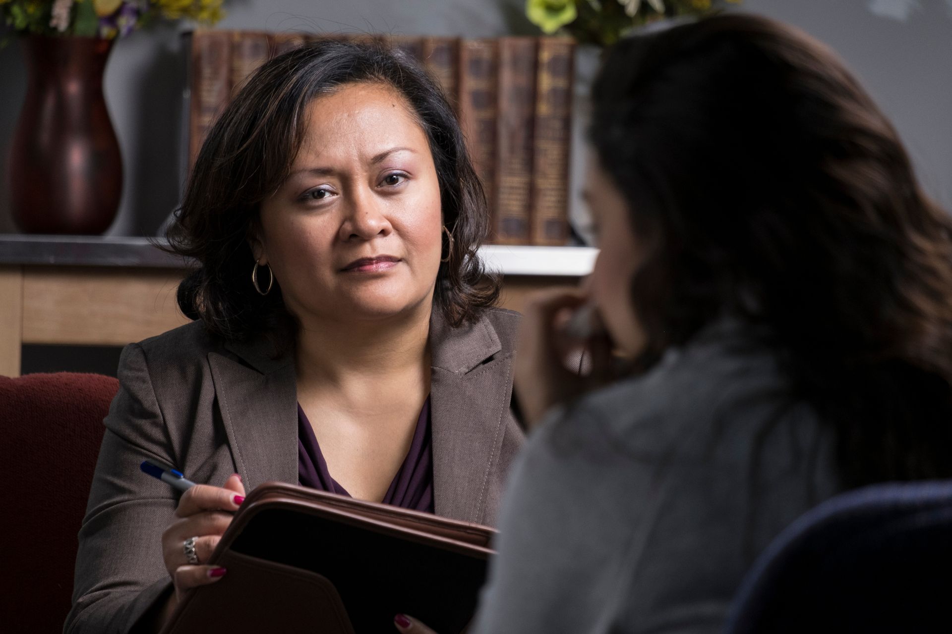 Two women in a quiet discussion, one holding a folder in a bookshelf-lined room.