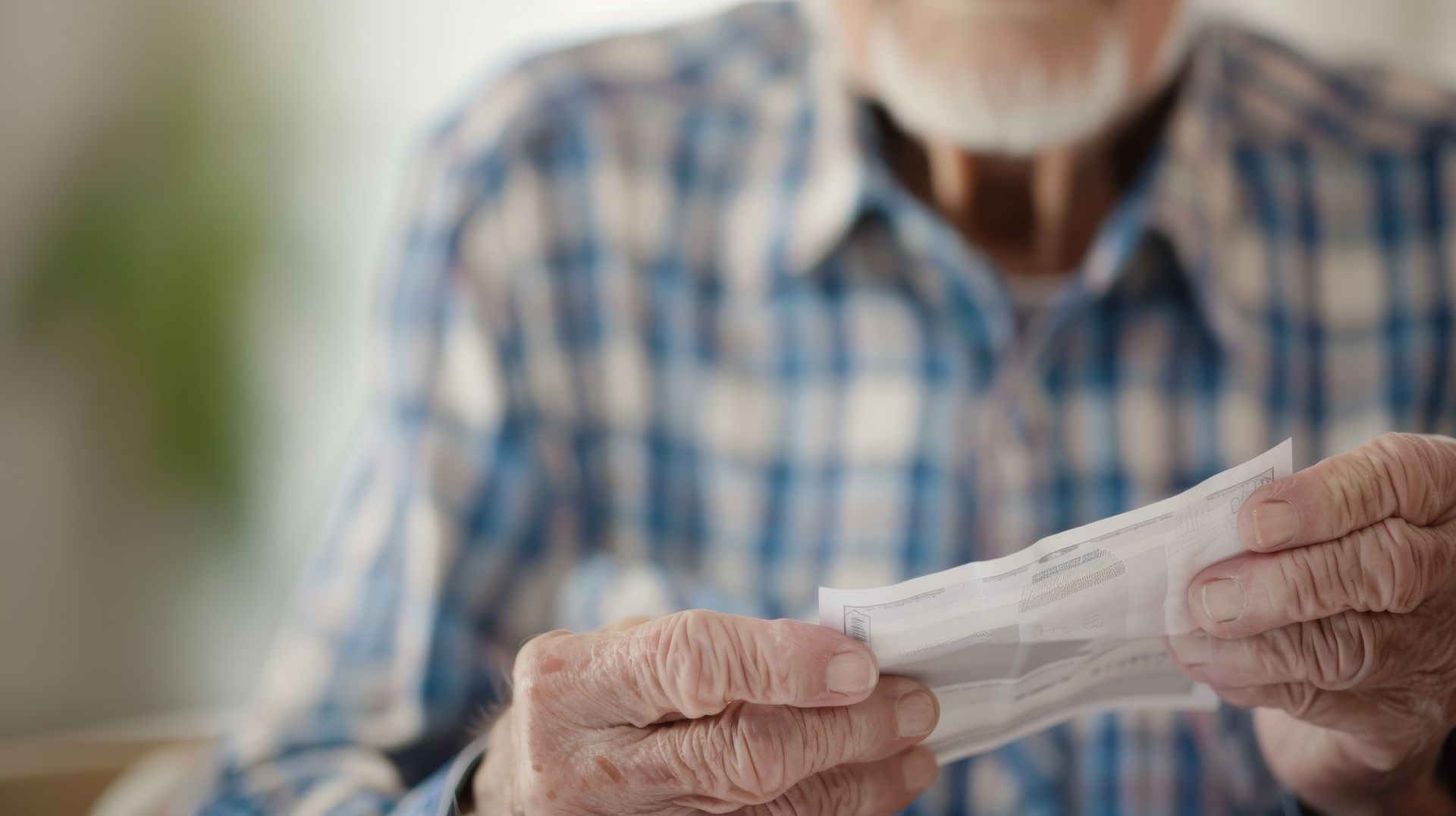 Hands holding a folded paper bill in a blue plaid shirt, with a blurred indoor background.