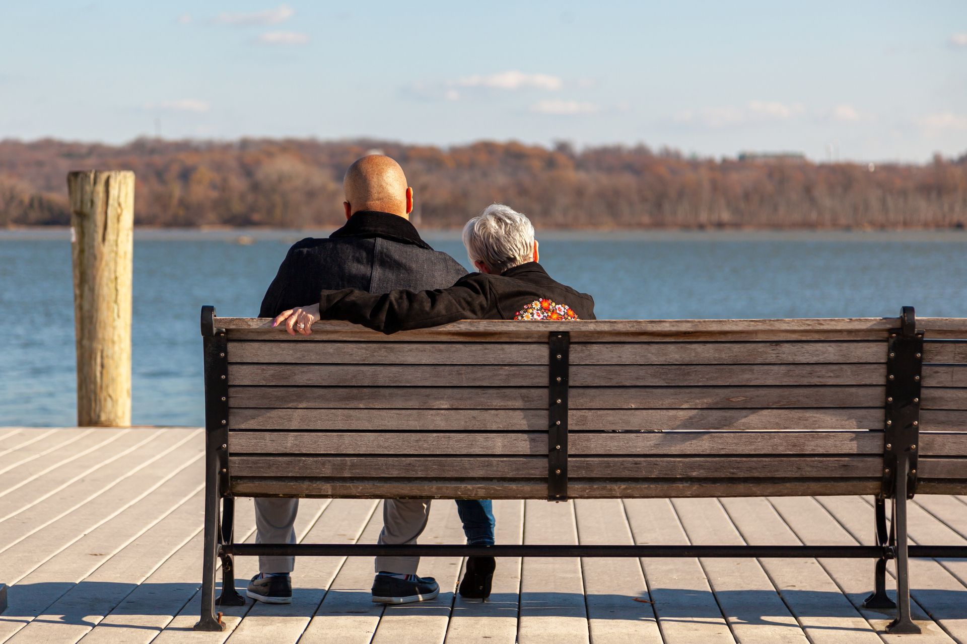 Two people sit on a bench facing a calm lake on a wooden dock