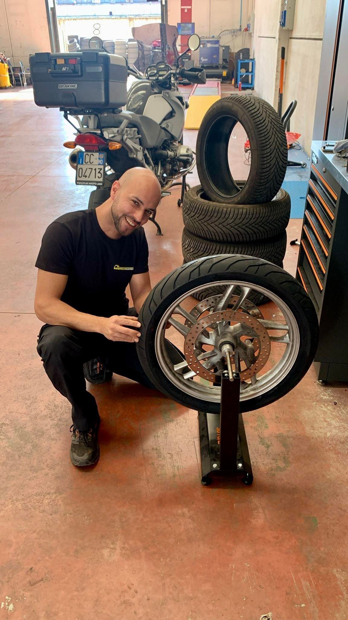 Un meccanico è inginocchiato accanto a una ruota di motocicletta in un'officina, con pneumatici impilati sullo sfondo.
