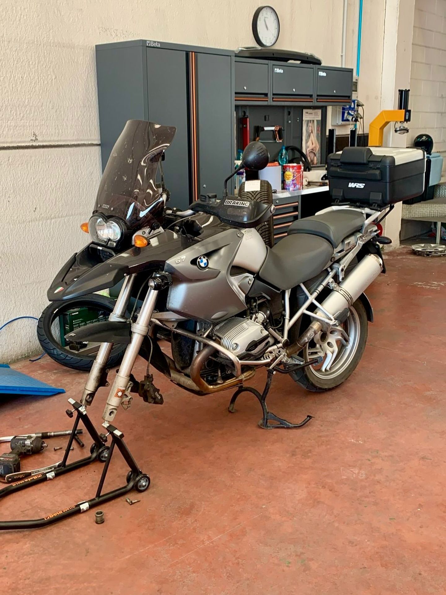 Una motocicletta argentata su un cavalletto in un garage, con armadietti e attrezzi sullo sfondo.