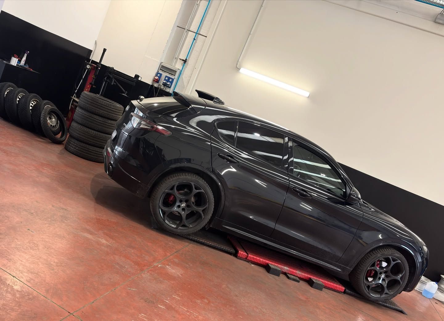 Una coupé sportiva nera in un garage, parcheggiata accanto a una pila di pneumatici da corsa su un pavimento rosso.