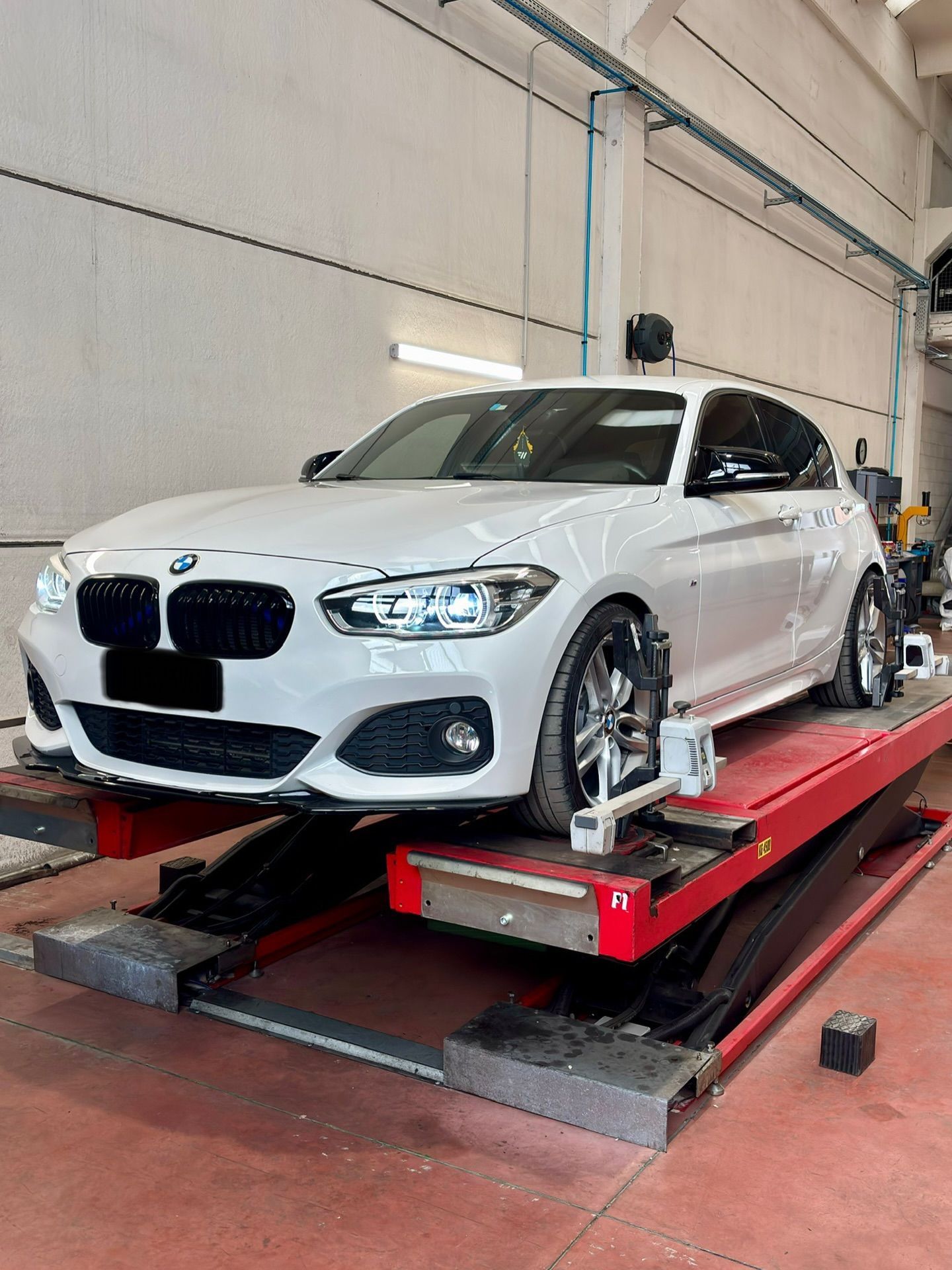 BMW bianca su un ponte sollevatore rosso in un'officina, ruote anteriori fissate con morsetti