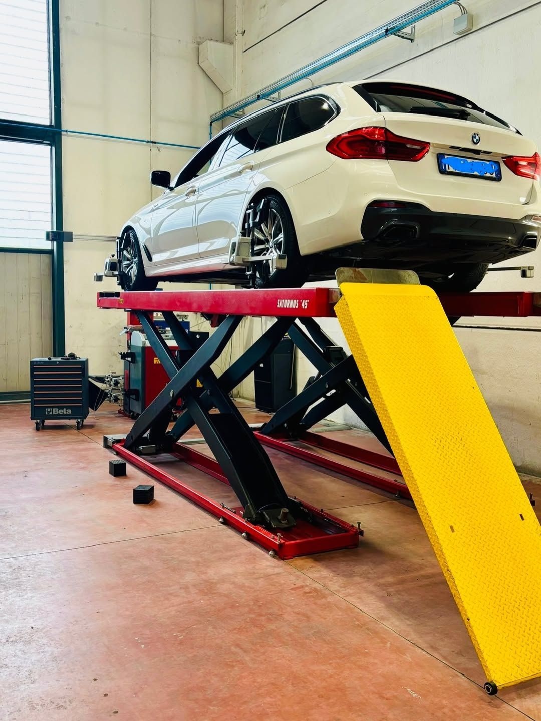 Un'auto bianca su un sollevatore idraulico rosso in un garage, con rampa gialla e pavimento arancione.