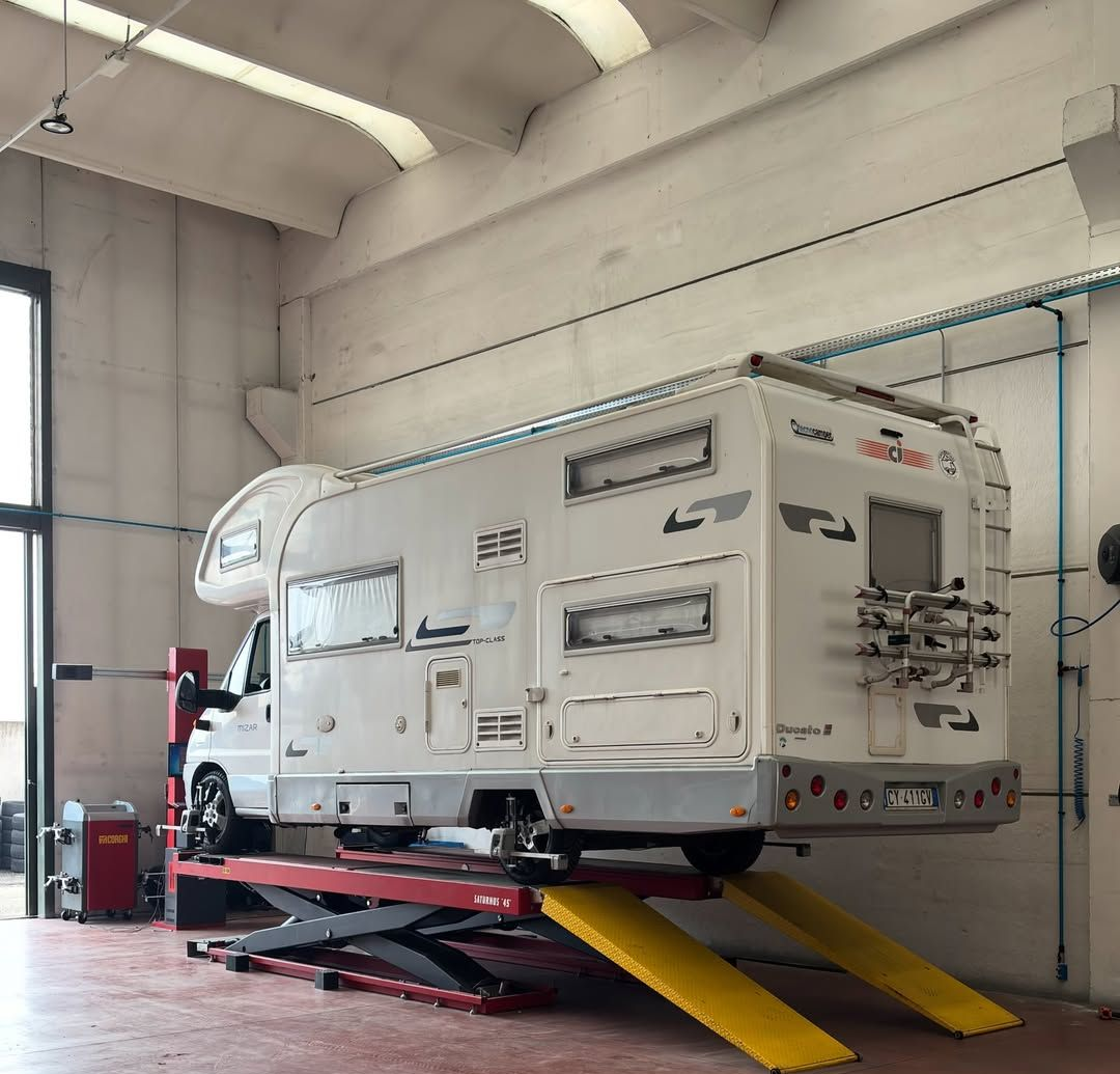 Camper bianco su un sollevatore idraulico all'interno di un'officina, con rampe gialle e attrezzature di supporto rosse.