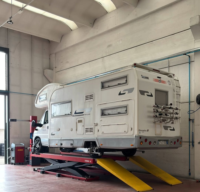 Camper bianco su un sollevatore idraulico all'interno di un'officina, con rampe gialle e attrezzature di supporto rosse.