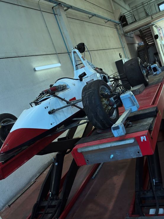 Auto da corsa bianca su un ponte sollevatore in un garage, con pannelli sottoscocca rossi e attrezzi nelle vicinanze.