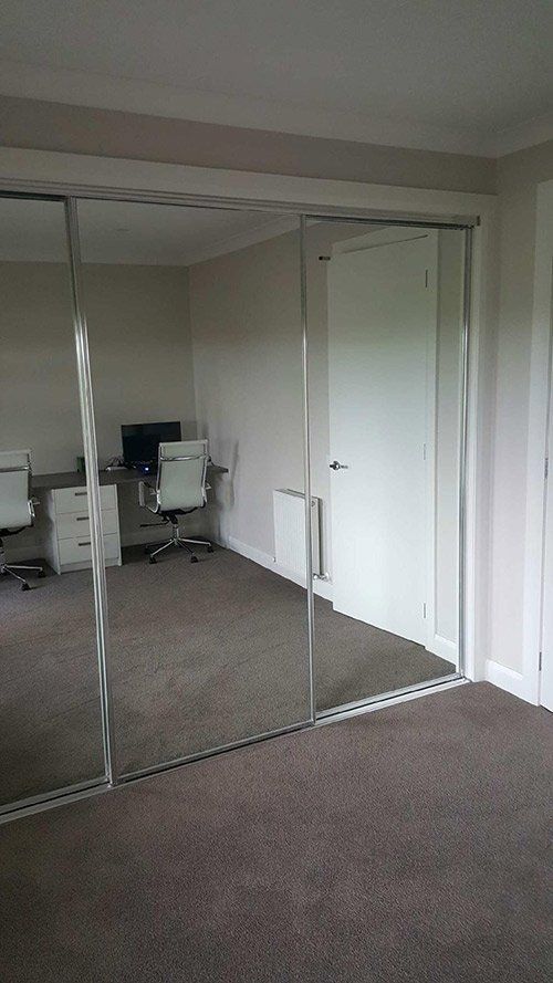 A room reflection in sliding mirrored closet doors, showing a desk with a computer and chair, and a white interior door.