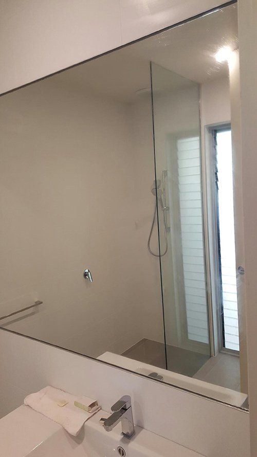 A modern bathroom vanity with a large mirror reflecting a glass-enclosed shower and a window with blinds.