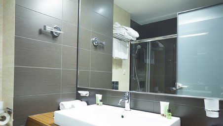 A modern bathroom featuring a white sink, large mirror, dark tiled walls, and towels neatly folded on a shelf.