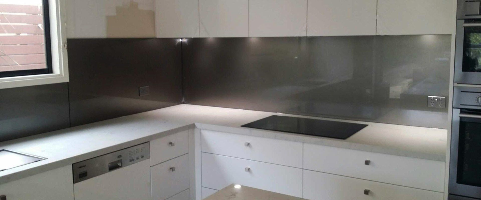 A modern kitchen featuring white cabinets, a light grey countertop, and a sleek, dark grey glass splashback.