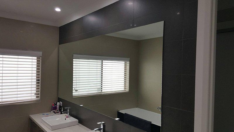 A modern bathroom featuring a wall-mounted rectangular mirror above a white sink with a window and horizontal blinds.