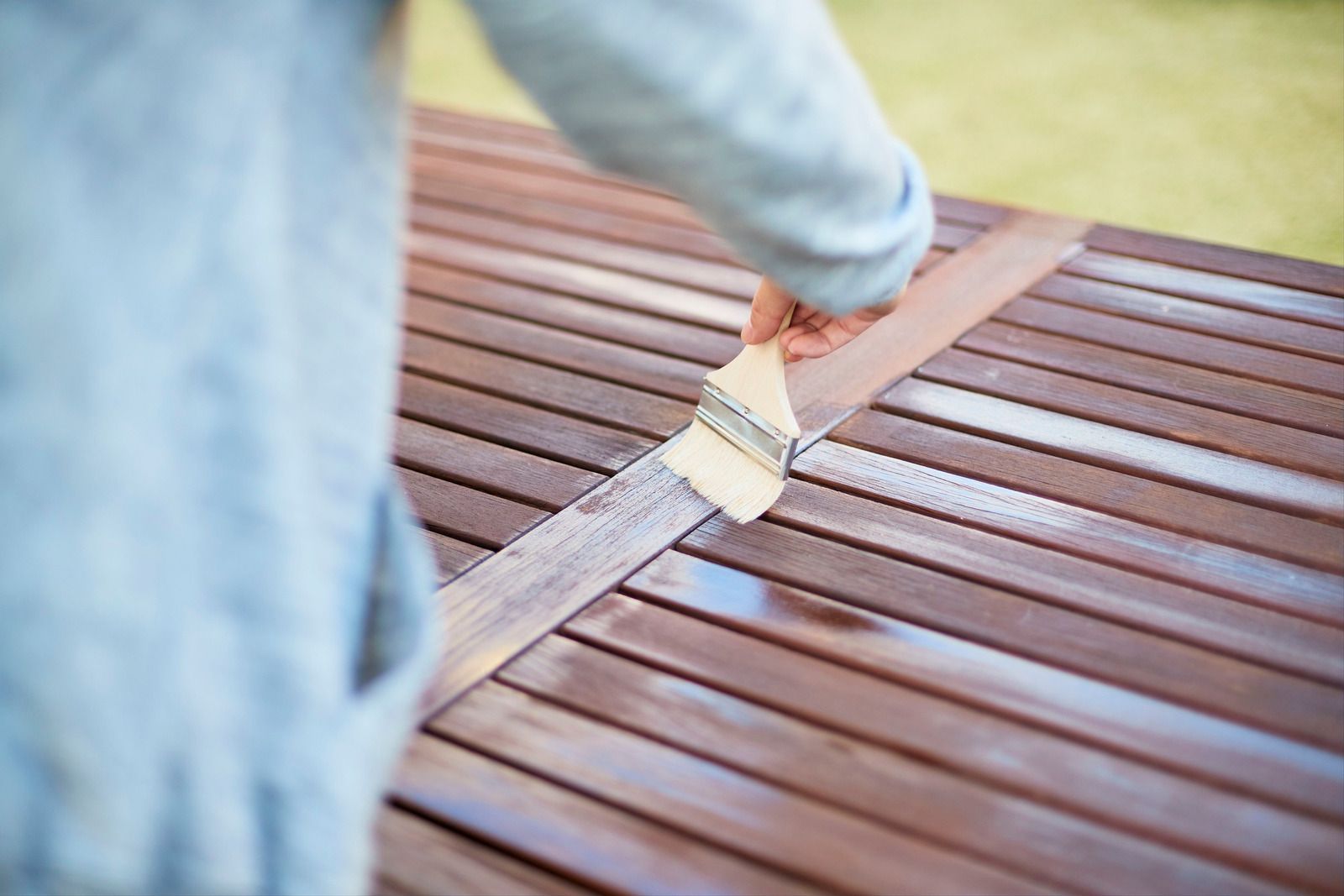 Hand using a paintbrush to apply brown paint to a wooden bench slat
