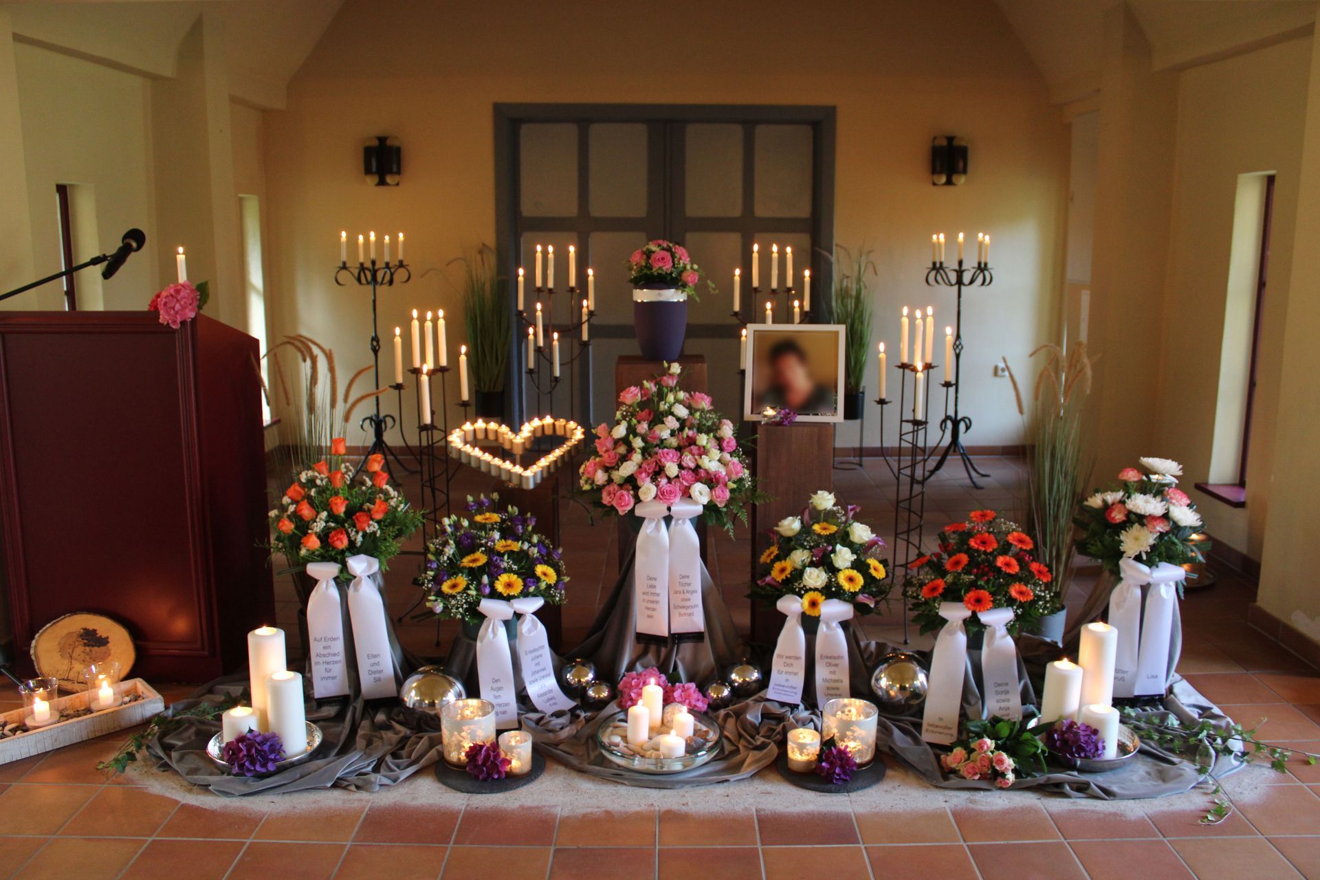 Trauerdekoration: Blumen, Kerzen, Foto, herzförmige Lichter; drinnen mit Rednerpult.