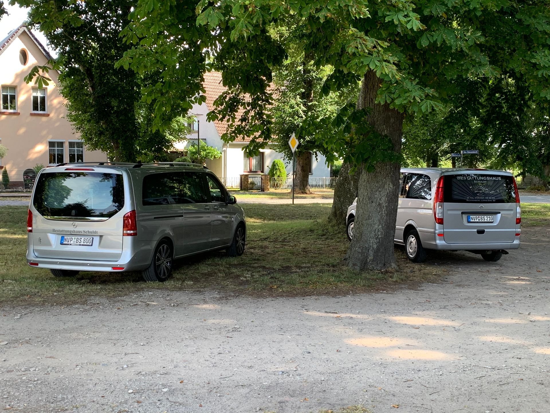 Zwei graue Lieferwagen parken unter Bäumen, im Hintergrund ist ein helles Gebäude zu sehen.