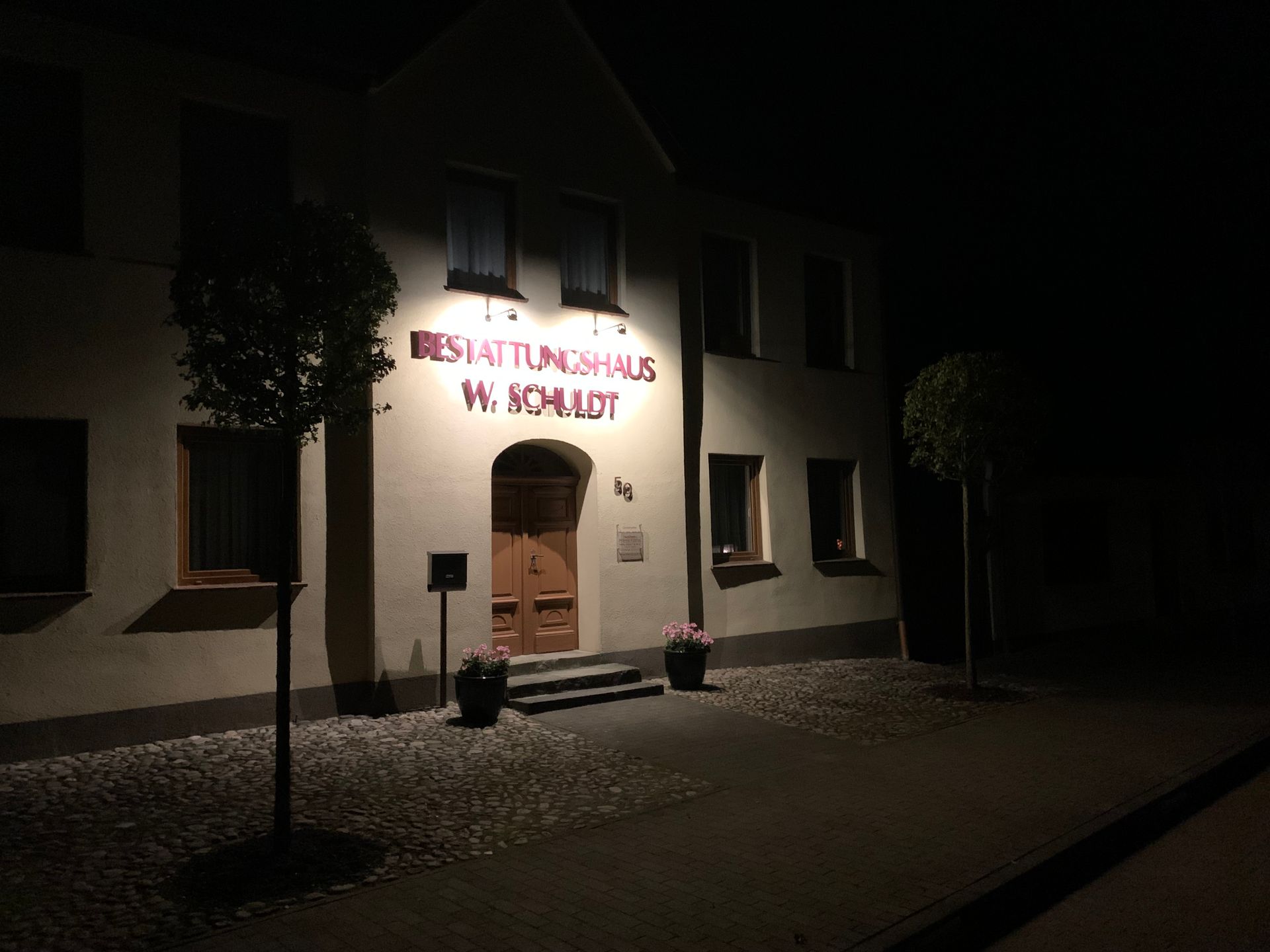 Gebäude des Bestattungshauses Schuldt bei Nacht mit beleuchtetem Schild „BESTATTUNGSHAUS W. SCHULDT“ und Tür.