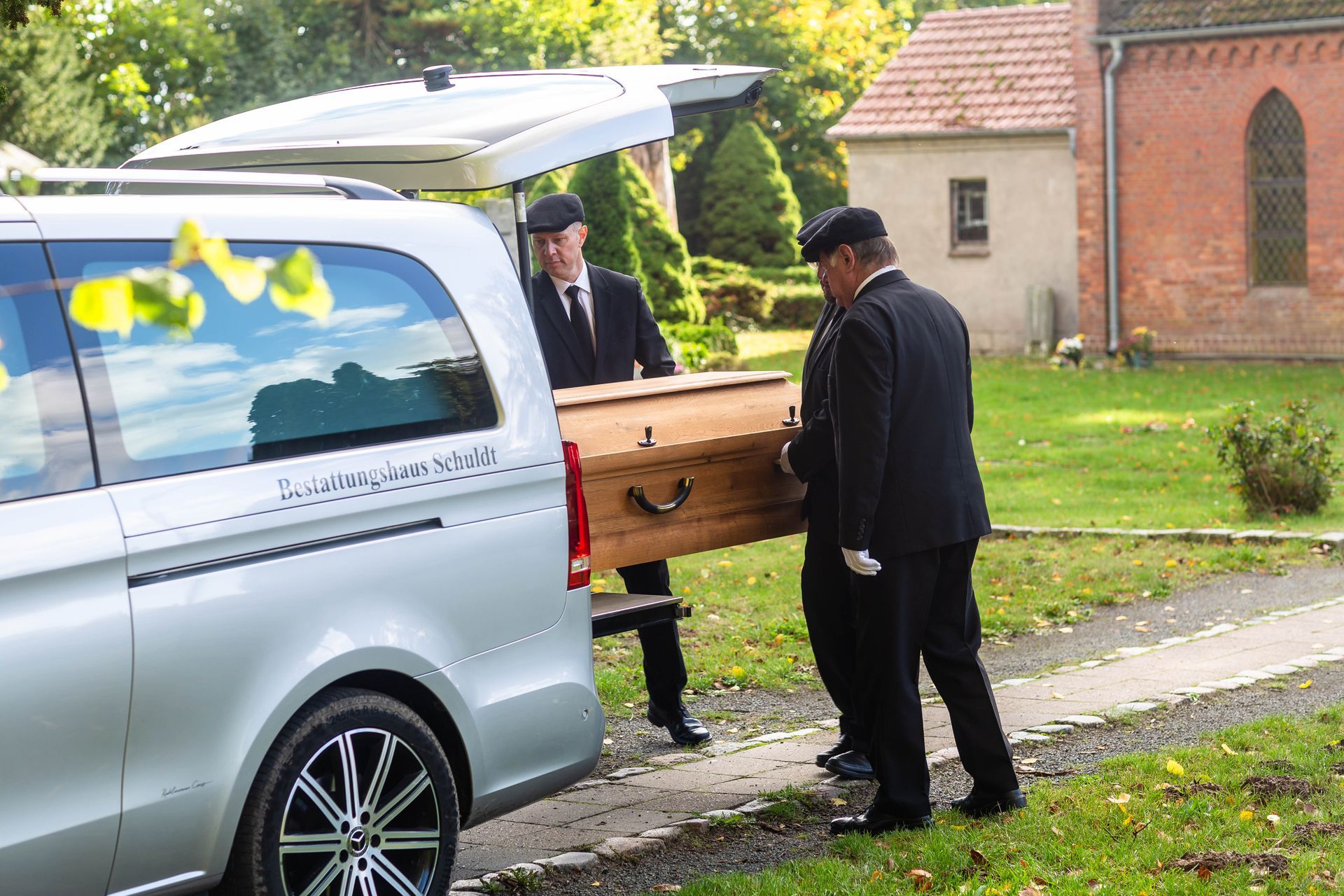 Zwei Männer in schwarzen Anzügen tragen einen Holzsarg aus einem silbernen Leichenwagen vor einer Kirche.