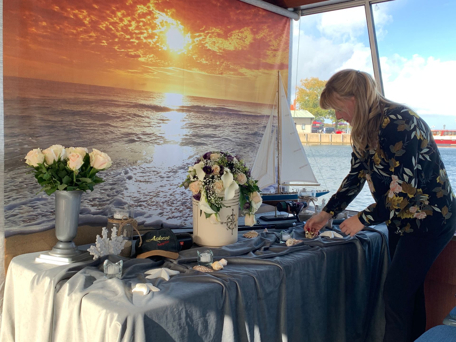 Frau dekoriert einen Tisch mit Blumen, Kuchen und Muscheln; im Hintergrund der Sonnenuntergang am Meer.