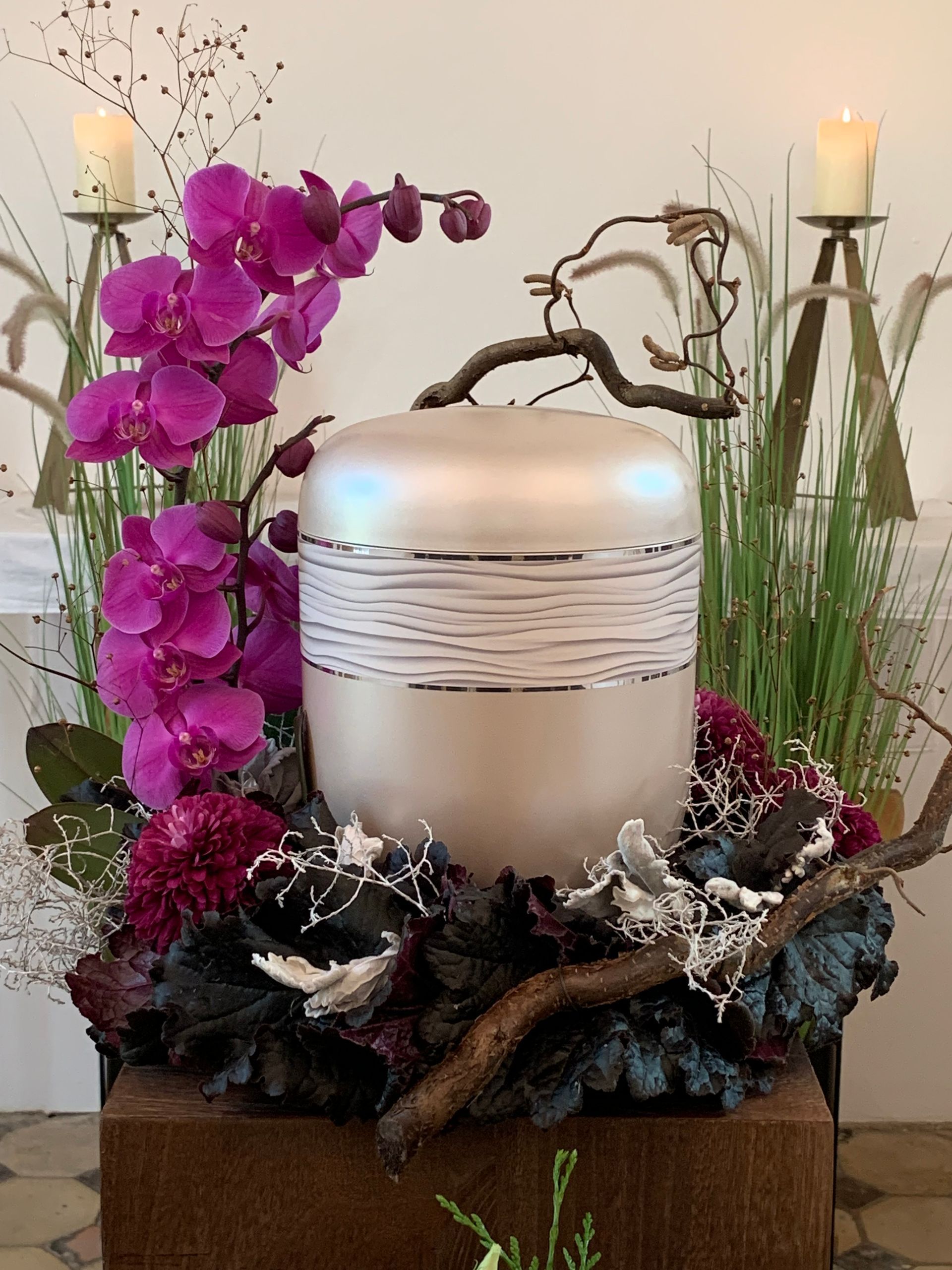 Urne, umgeben von violetten Orchideen, Kerzen und Grünpflanzen, auf einem braunen Ständer.