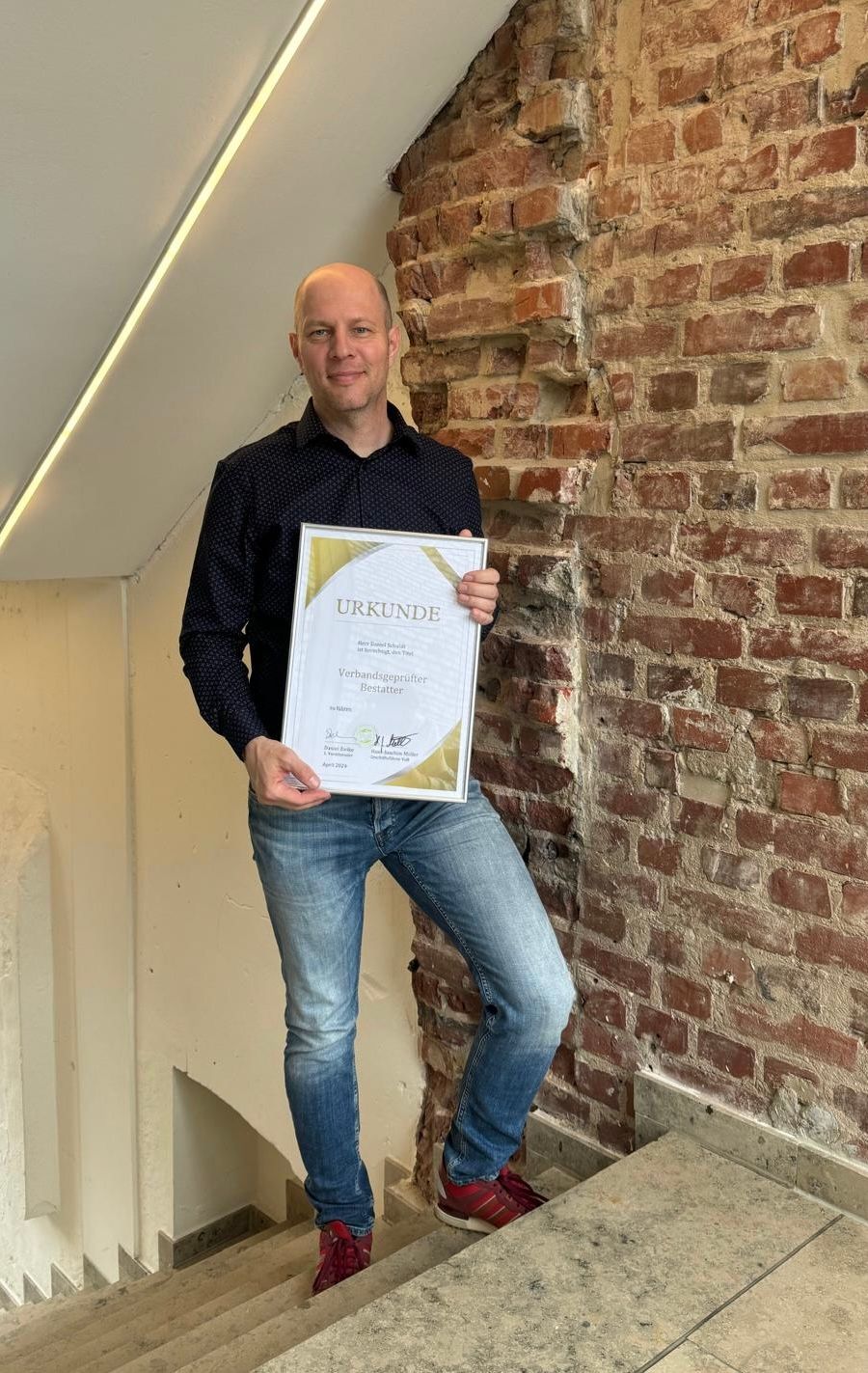Bestatter Daniel Schuldt steht stolz mit seiner Urkunde als verbandsgeprüfter Besatter in der Hand auf einer Treppe.
