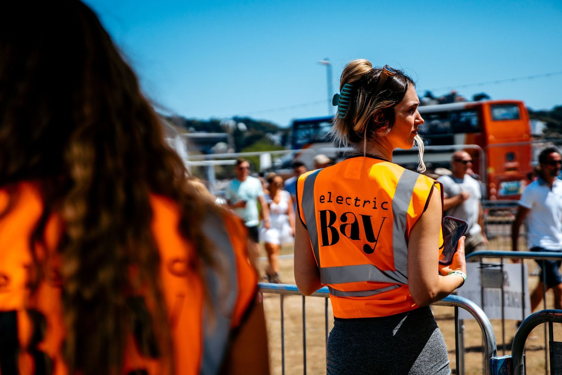 A woman wearing an orange vest and headphones is standing in front of a fence.