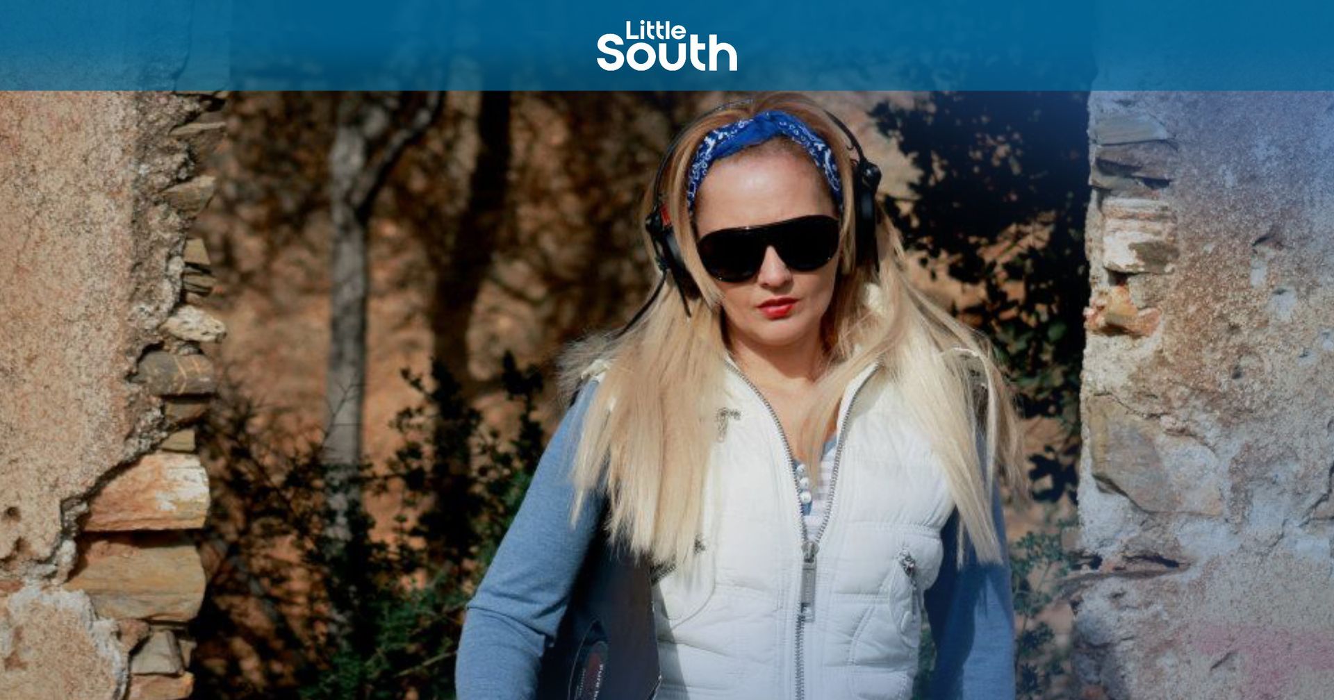 A woman wearing headphones and sunglasses is standing in front of a stone wall.