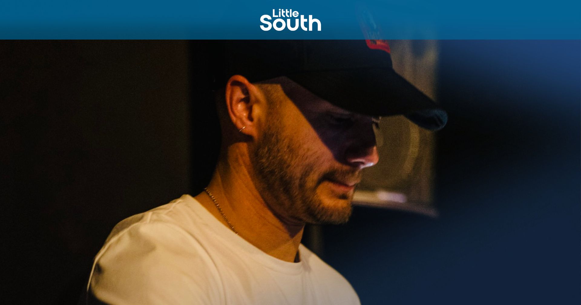 A man wearing a hat and a white shirt is sitting in a dark room.