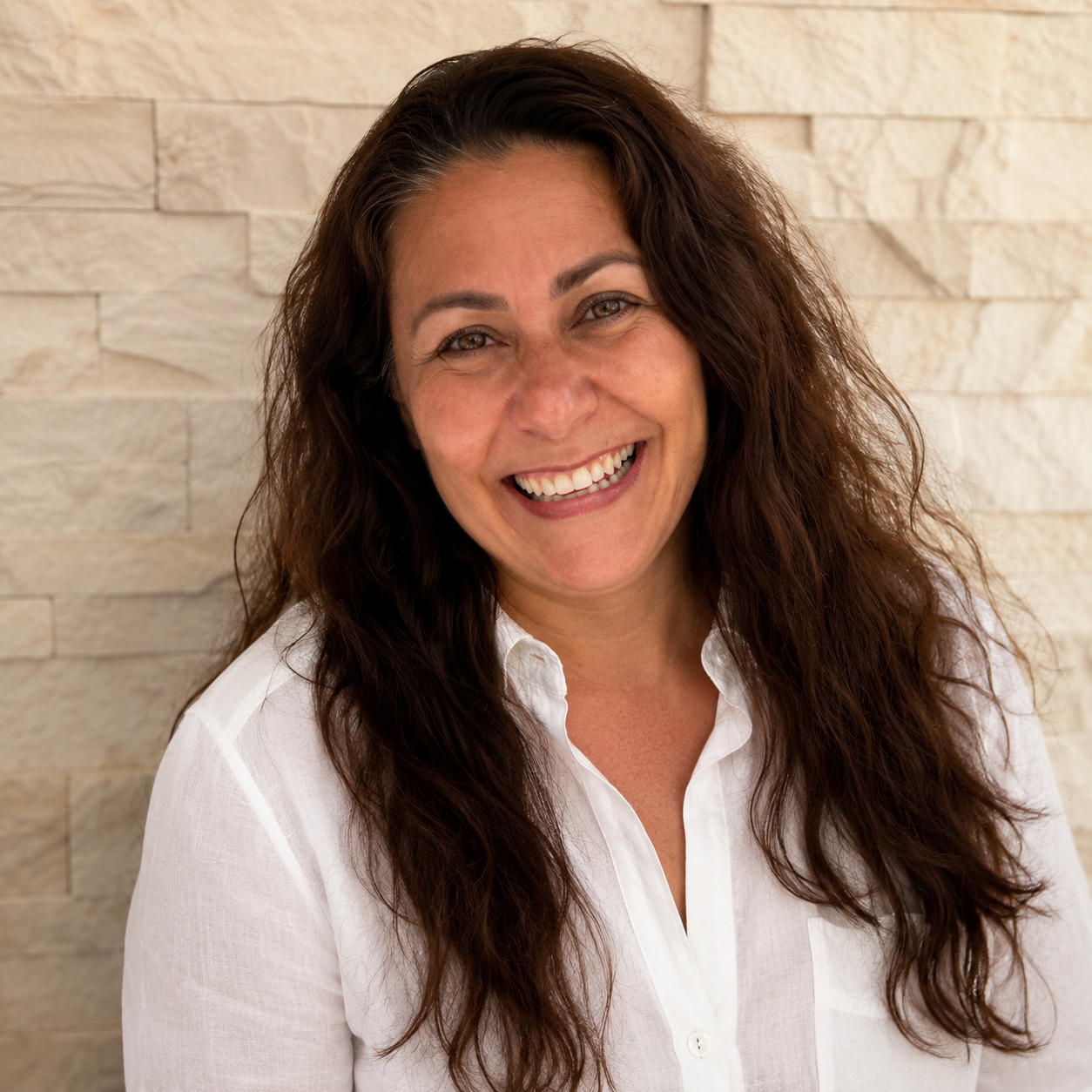 Mujer sonriente con camisa blanca de pie contra una pared de piedra beige.