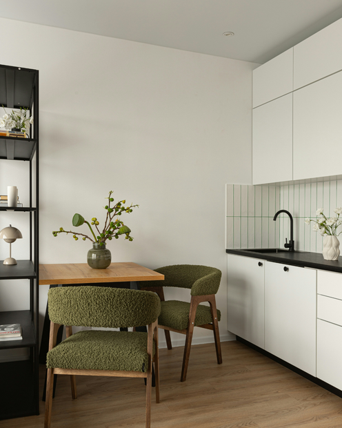 Rincón moderno de cocina-comedor con mesa de madera, sillas verdes, armarios blancos y encimera negra.