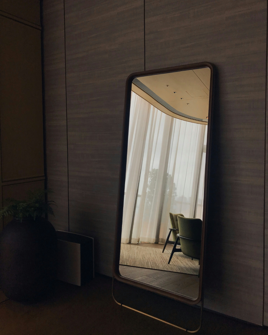 Un espejo alto de pie, apoyado contra la pared, refleja una habitación luminosa con cortinas transparentes y una silla verde.