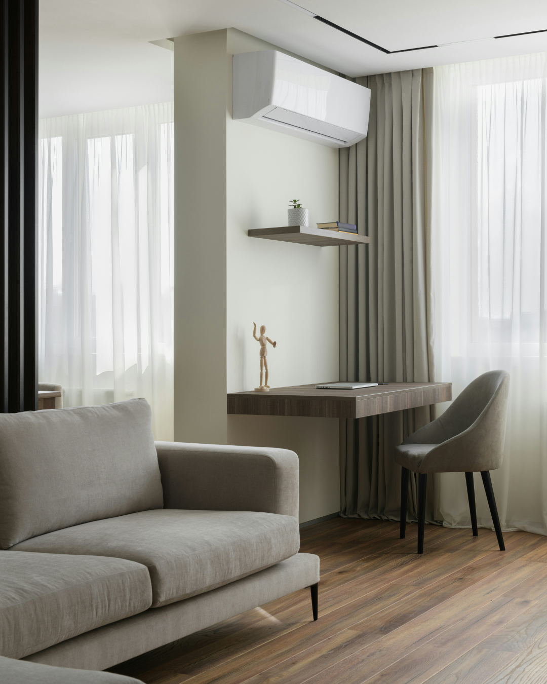Sala de estar moderna con sof&aacute; gris, rinc&oacute;n de escritorio, silla y grandes ventanas con cortinas transparentes.