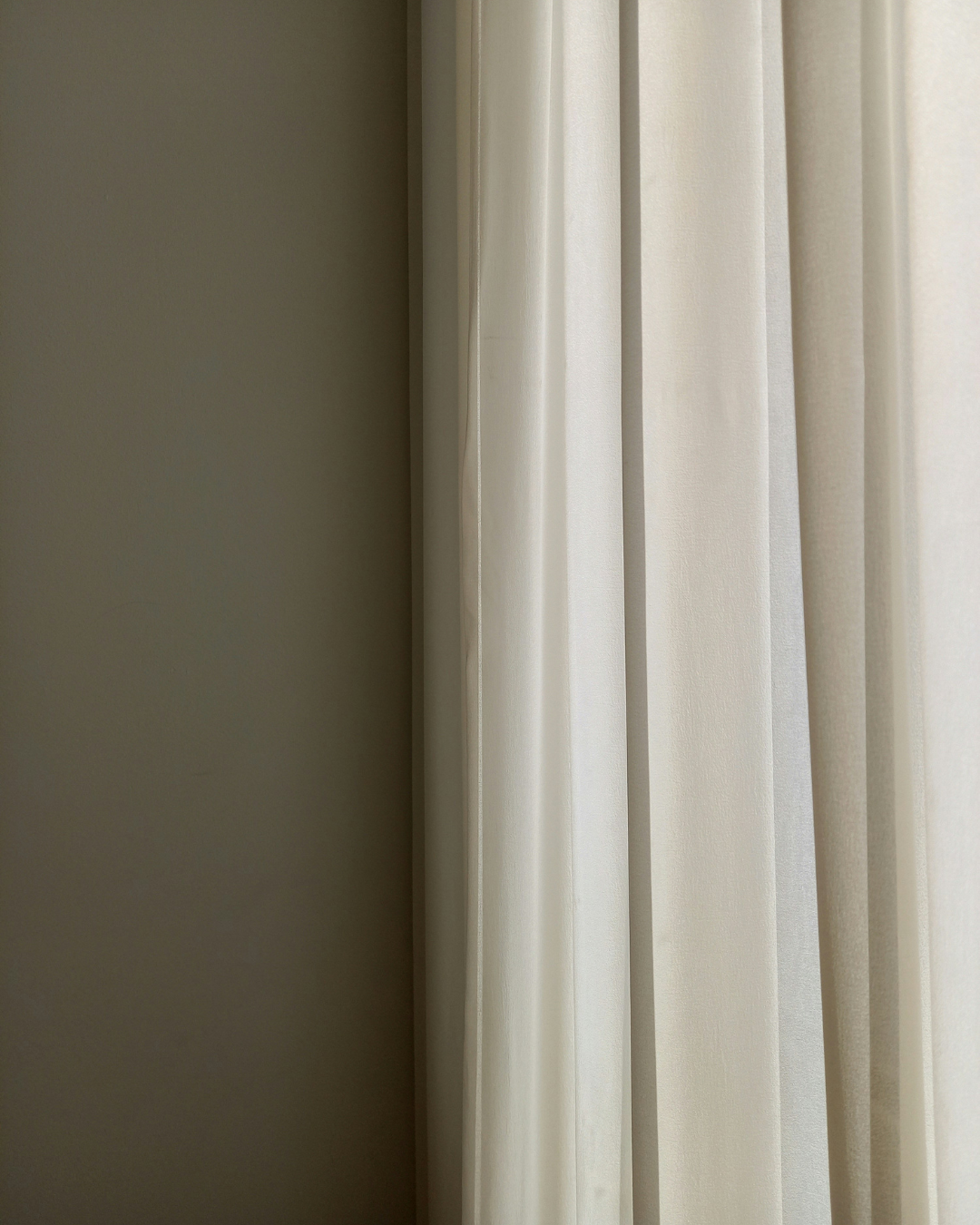 Cortinas de color beige cuelgan junto a una pared oscura, suavemente iluminada por luz natural.