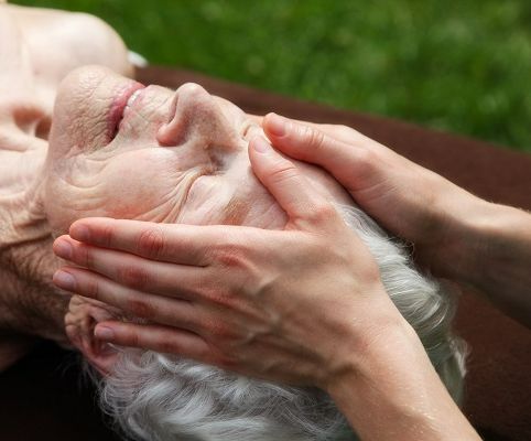Handen die een gezichtsbehandeling geven aan een oudere vrouw voor schoonheidsverzorging op maat van Maatjeshulp.