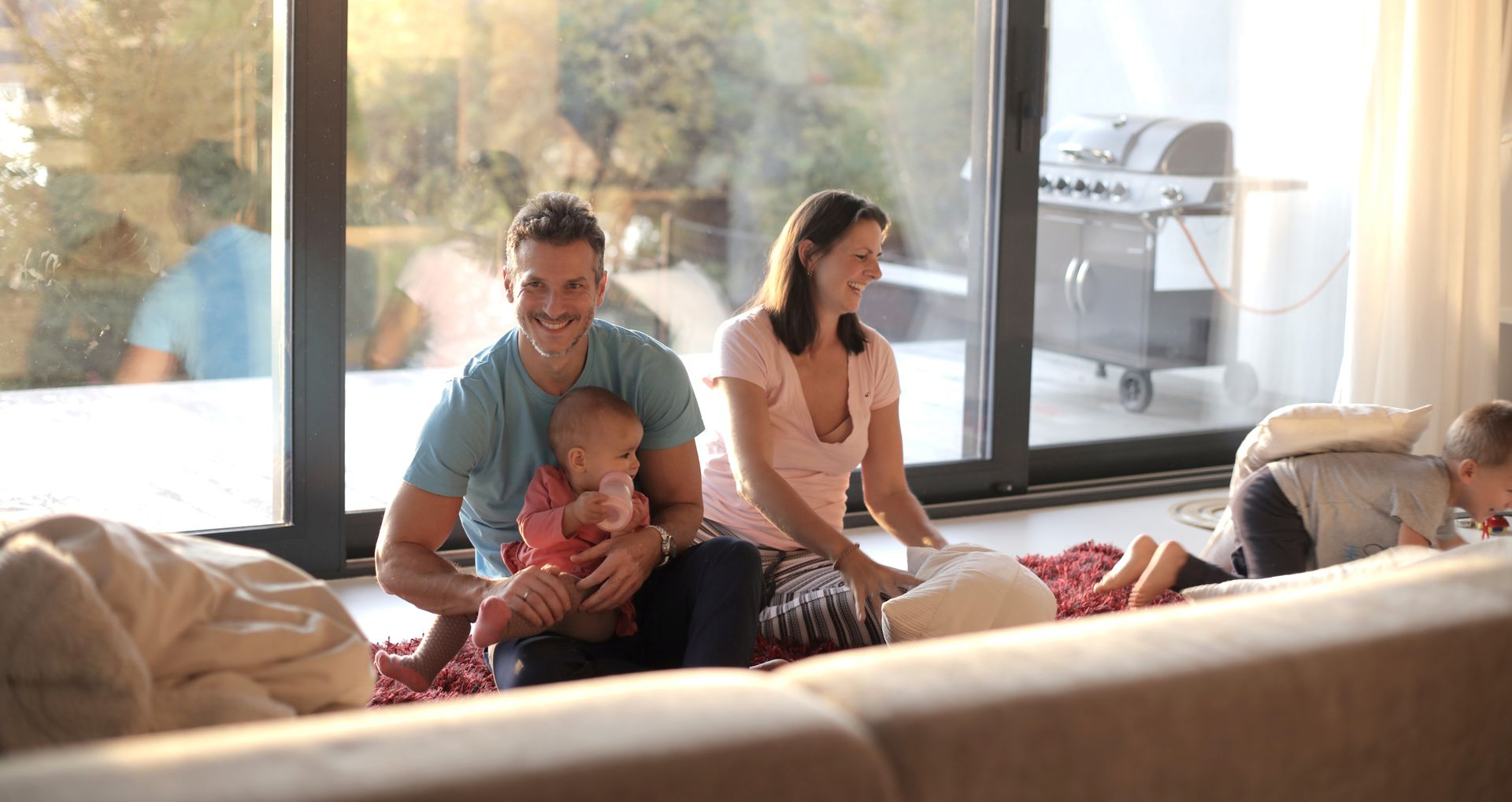 Family relaxing on a couch in a sunlit living room, with a man holding a baby and a woman sitting nearby.