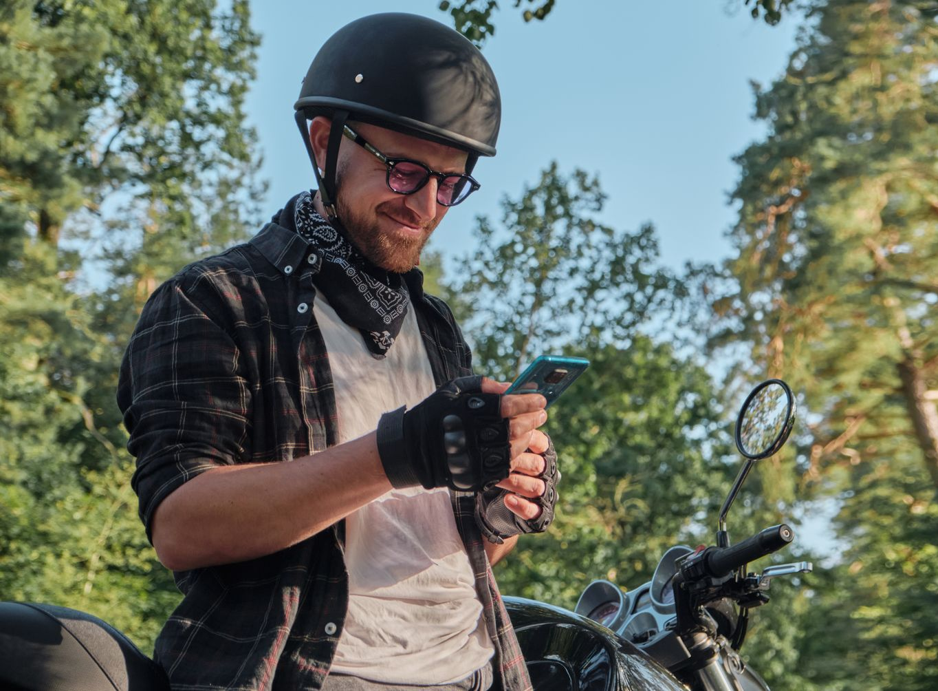 Man in a helmet checking his phone beside a motorcycle in a wooded area