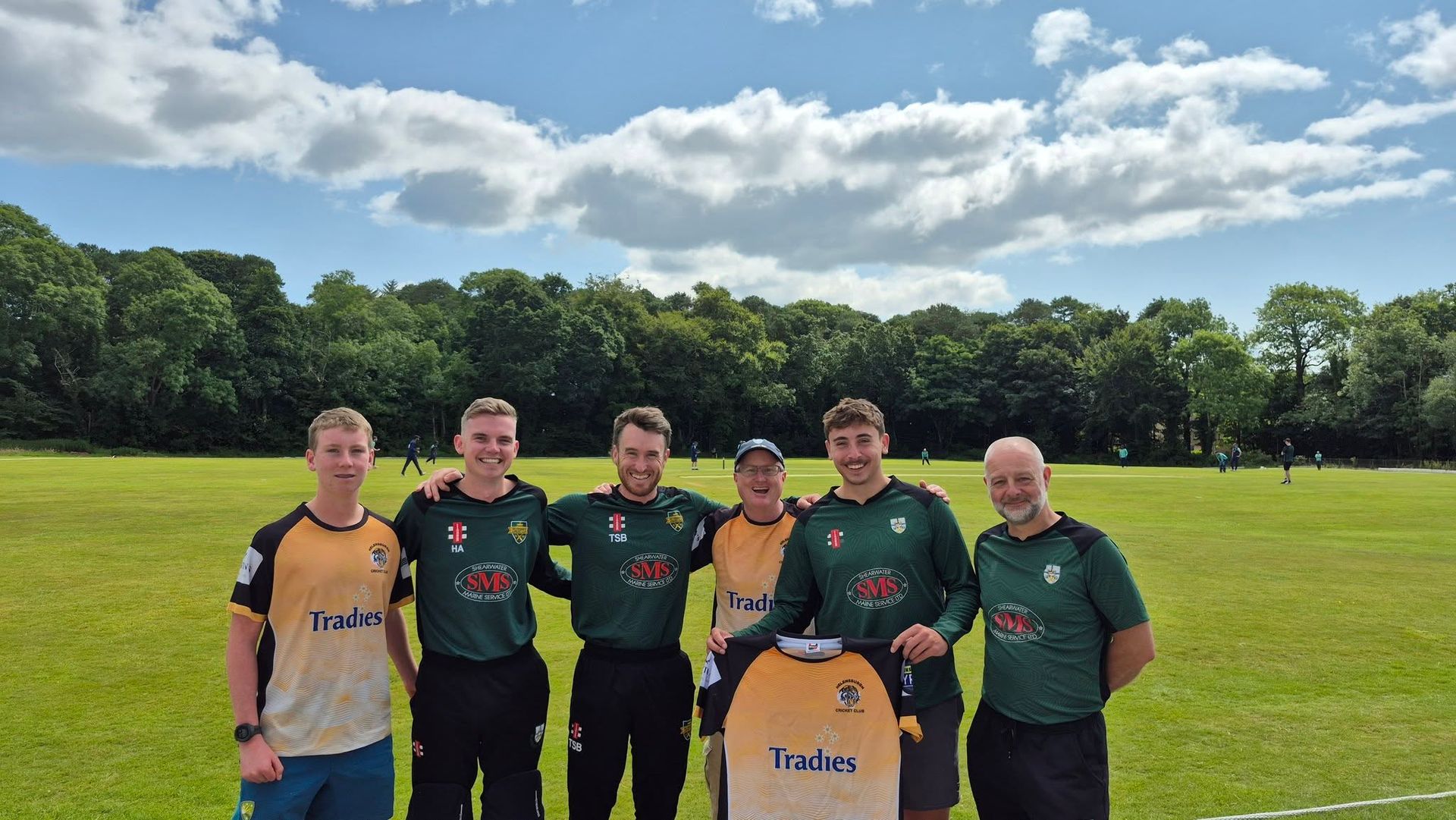 Six soccer players pose on a sunny field with trees behind them, holding a yellow jersey.