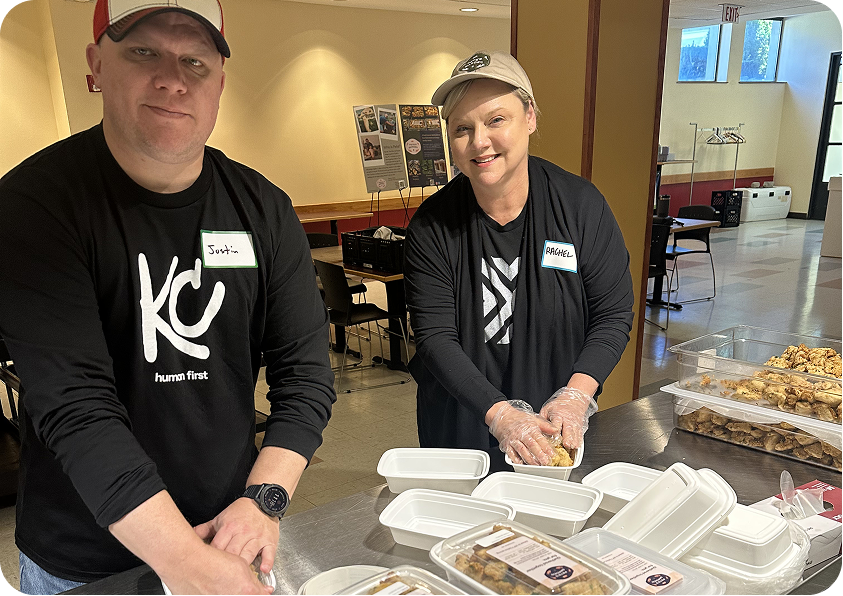 Two volunteers in matching black long-sleeved shirts and hats pack food containers in a brightly lit room.