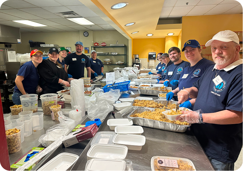 A group of volunteers in dark blue shirts packing meals into containers in an industrial kitchen.